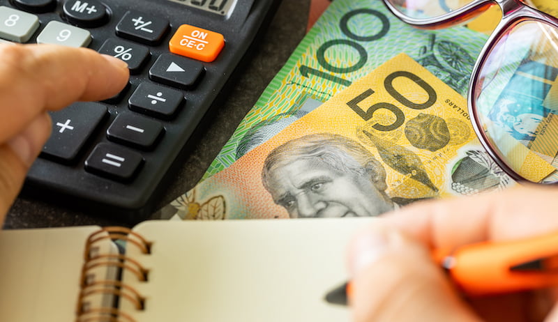 Australian money beside a calculator and hand holding a pen in front of a notepad